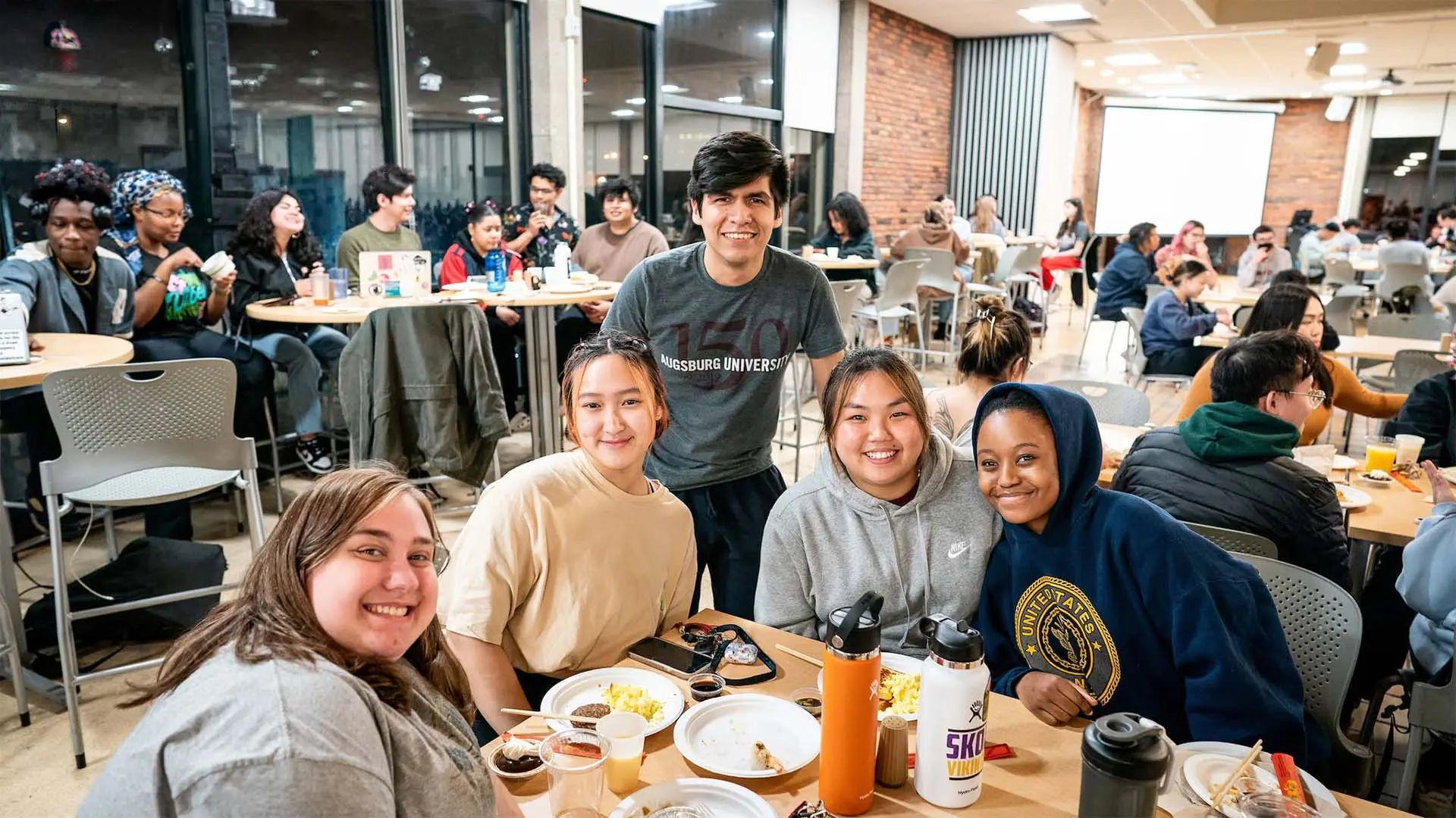 Five students smile together while sharing a meal in a busy campus dining hall.