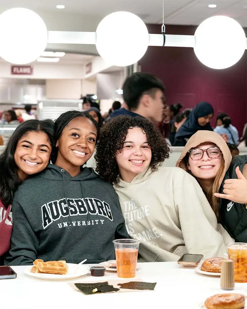 Four students smile together while enjoying breakfast in a busy campus dining hall.